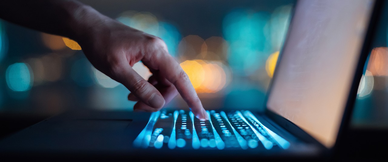 Close up of woman's hand typing on computer keyboard in the dark against colourful bokeh in background, working late on laptop at home